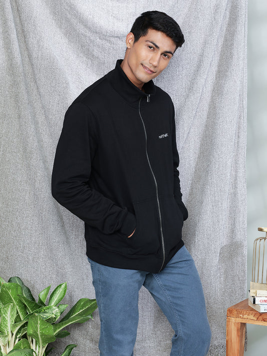 A person wearing the Black Beauty High Neck Jacket made of 100% organic cotton from Northmist, with hands in the pockets, stands in front of a gray curtain backdrop. A green potted plant is on the left side and a wooden table with a birdcage is on the right. The person is smiling and looking at the camera.
