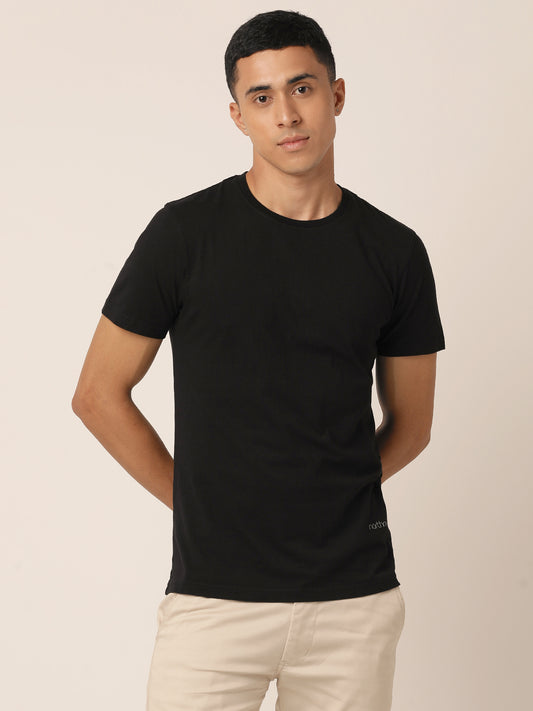 A person with short dark hair is wearing a Bare Black Crew Neck T-Shirt by Northmist made from 100% Organic Cotton and light beige pants. They are standing against a neutral background with their hands behind their back.