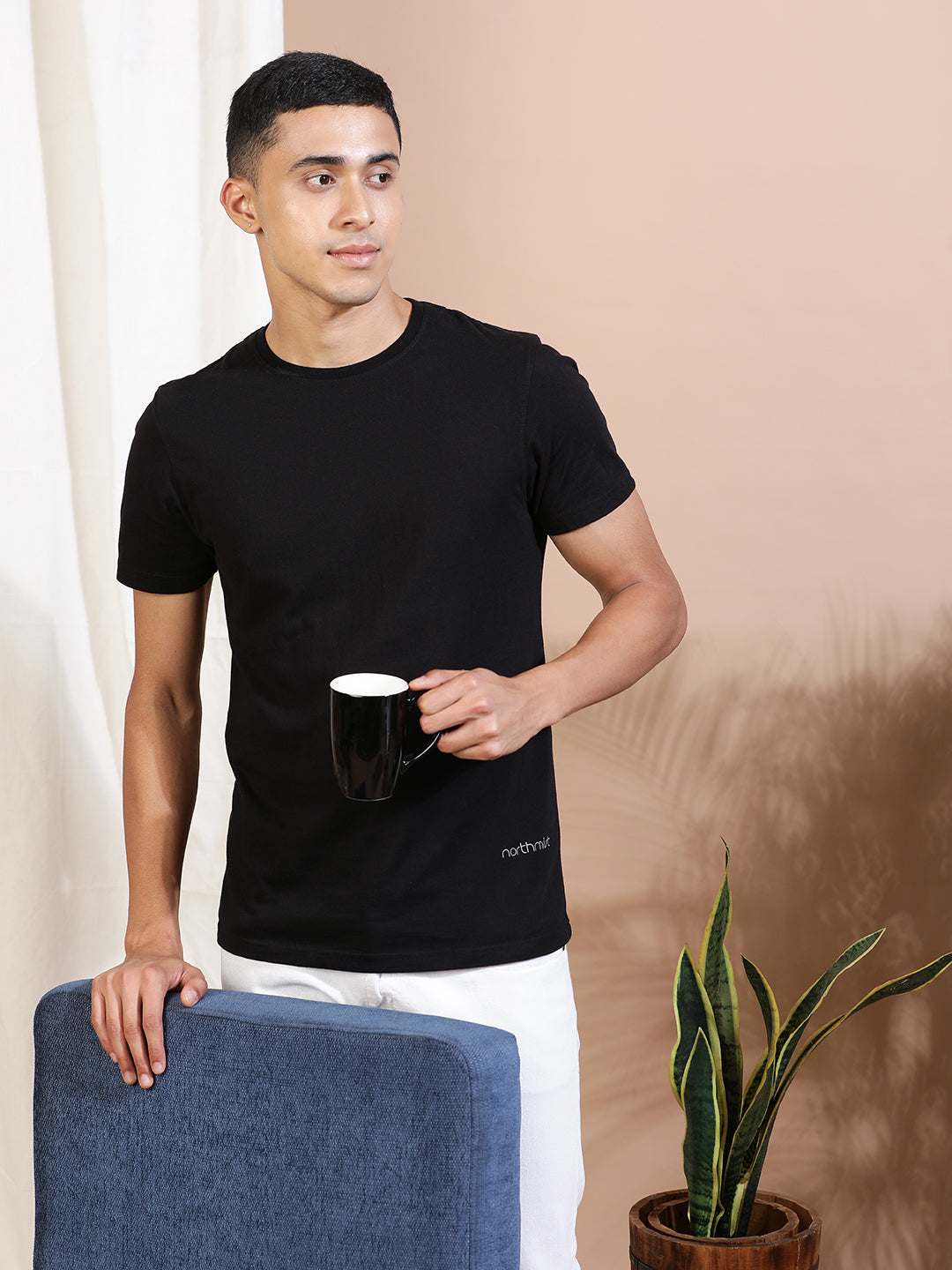 A man in a Bare Black Crew Neck T-Shirt by Northmist and white pants stands next to a blue chair while holding a black mug. He is looking off to the side. A plant in a pot is placed beside the chair, and a curtain partially covers the beige wall in the background. The shirt is made from 100% organic cotton.