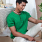 A man wearing a green Abundant Green Henley T-Shirt by Northmist and white pants sits on the floor next to a table covered with a patterned cloth. On the table are a lantern, some books, and a plant. The man is looking down with a relaxed expression, and an abundant green plant is visible behind him.