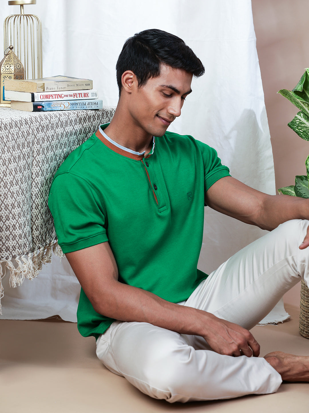 A man wearing a green Abundant Green Henley T-Shirt by Northmist and white pants sits on the floor next to a table covered with a patterned cloth. On the table are a lantern, some books, and a plant. The man is looking down with a relaxed expression, and an abundant green plant is visible behind him.