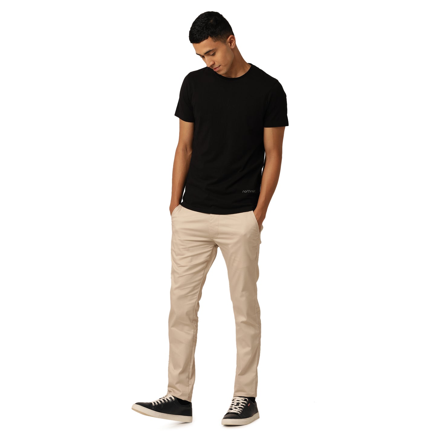 A happy guy stands against a plain white background, wearing a black t-shirt from the Alpha Combo Crew Neck T-Shirts (Pack of 3) by Northmist, beige pants, and black sneakers with white soles. He has his hands in his pockets and is looking down slightly with a relaxed posture.