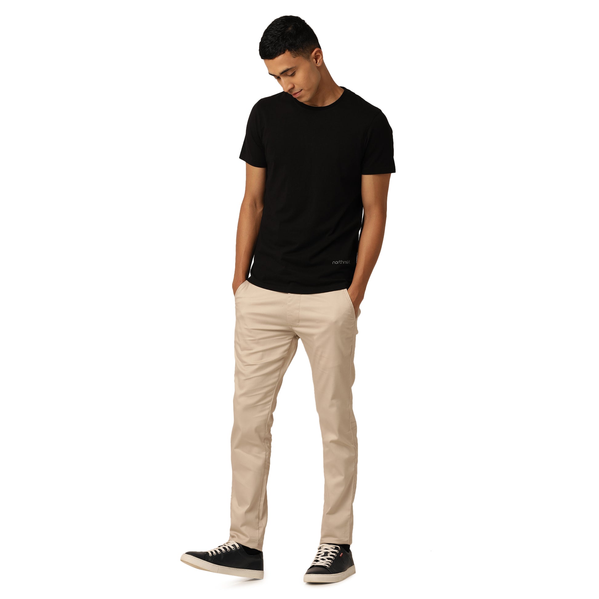 A happy guy stands against a plain white background, wearing a black t-shirt from the Alpha Combo Crew Neck T-Shirts (Pack of 3) by Northmist, beige pants, and black sneakers with white soles. He has his hands in his pockets and is looking down slightly with a relaxed posture.
