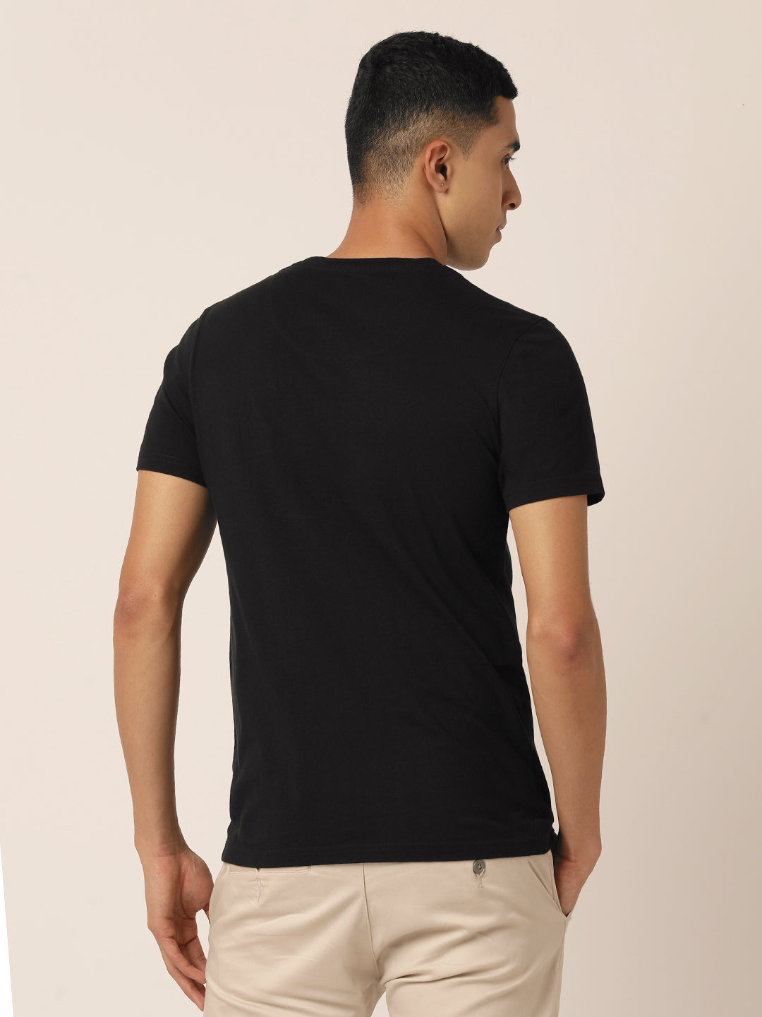 A person with short dark hair is shown from behind, wearing a Northmist Bare Black Crew Neck T-Shirt made from 100% organic cotton and beige pants. The background is neutral and light-colored. The person is turned slightly to the left.