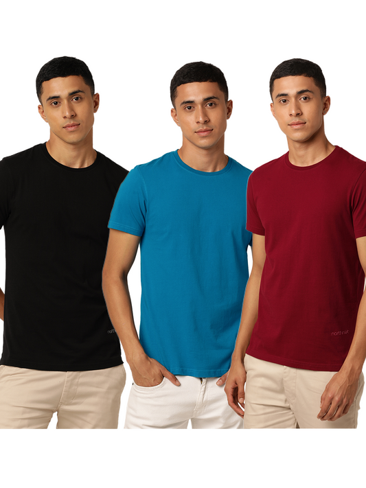 Three identical, happy guys with short black hair wear different colored Alpha Combo Crew Neck T-Shirts (black, blue, and red) in organic cotton and beige pants. They stand side-by-side against a plain white background. The T-shirts are part of a pack of 3 from Northmist.