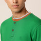 A person with short hair wearing an organic cotton, Abundant Green Henley T-Shirt by Northmist featuring buttons and a brown and grey trimmed collar. The individual is looking slightly to the left against a plain background. The shirt has a small emblem on the left chest.