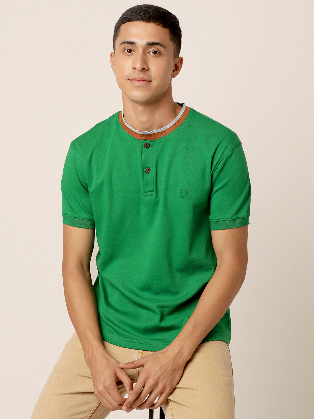A young man wearing a Northmist Abundant Green Henley T-Shirt with buttons, light brown pants, and a simple necklace sits on a stool. The background is plain and light-colored, making the man the focal point of the image.